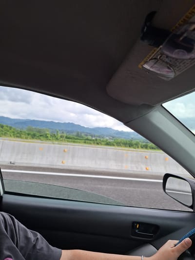 The image shows a view from inside a car traveling on a highway. The passenger is looking out the window at a landscape of green trees and distant mountains under a cloudy sky. A concrete barrier with orange markers lines the side of the highway. The car's interior is visible, including a portion of the dashboard, the driver's arm on the steering wheel, and a sun visor with glasses clipped to it. The location is specified as being in the community of Chibati, Georgia.