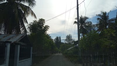 The image depicts a street scene in Pusong, Indonesia, under a cloudy sky. A paved road runs through the center, flanked by lush green vegetation and palm trees on both sides. A utility pole with numerous wires is visible on the right side of the road, a characteristic feature of the area. On the left, a small structure with a corrugated metal roof and a fence is present. Further down the road, two individuals are visible, one standing and another on a motorcycle, suggesting a rural or semi-urban setting. The overall atmosphere is serene, with a sense of everyday life in a tropical environment.