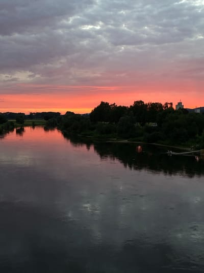 The image depicts a river scene at dusk in Rinteln, Germany. The sky is filled with dramatic clouds, transitioning from a vibrant pink and orange near the horizon to a darker gray and white at the top. A lush, dark green tree line borders the far bank of the river. On the right bank, a wooden pier extends into the water, with several individuals visible sitting or standing on it. Further in the distance, a building structure with a tower and what appears to be a smokestack is visible behind the trees. The river's surface is calm and reflects the colors of the sky and the surrounding trees, creating a mirror-like effect. The overall atmosphere is serene and picturesque, capturing the beauty of the natural landscape during twilight.