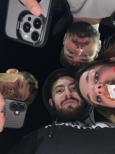 The image depicts four individuals in close proximity, looking downwards towards the camera, against a uniformly dark, non-descript background. This scene is located in Frătești, Romania.
**Subjects:**
1. **Upper-center:** An adult male with a dark beard and mustache, short dark hair, wearing a light-colored shirt. He gazes directly towards the camera.
2. **Right:** An adult female with long dark hair and dark eyes. She is holding a food item, partially wrapped in white paper with red text, near her mouth, appearing to be eating or about to eat.
3. **Lower-center:** An adult male with a full dark beard, mustache, and dark hair. He wears a black hooded sweatshirt with a white "NORTH" logo (likely "The North Face") and maintains a direct, neutral gaze towards the camera.
4. **Lower-left:** A young adult male with lighter, wavy hair and a clean-shaven face. His left ear is clearly visible. He looks towards the camera.
**Objects and Actions:**
* In the upper-left, a hand holds an iPhone (triple camera module, likely 13 Pro/14 Pro series) in a clear case with "BRACKET" branding, oriented with its cameras facing the viewer.
* In the lower-left, another hand holds a second iPhone (triple camera module) in a dark, ribbed case, also oriented with its cameras facing the viewer.
* The overall composition suggests a group selfie or a close-up group portrait taken from a low angle, likely at night or in a very dimly lit indoor environment due to the lack of visible background details.