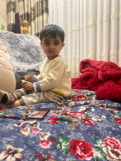 An indoor scene in Galaosiyo, Uzbekistan, shows a young child seated on a dark blue floral patterned bed or floor covering. The child has dark hair and eyes, wears a light yellow long-sleeved top with blue cuffs, and light-colored pants patterned with what appears to be a "Gucci" logo. The child holds a small green object in their right hand and looks directly at the camera. To the left, part of an adult's lower body, clothed in light fabric and patterned socks, is visible. Behind the child, a grey and white patterned fabric covers a surface, and a gold-colored decorative object rests on it. To the child's right, a large red fluffy object, likely a blanket or pillow, is present. In the foreground, a black smartphone lies on the floral covering, displaying an image of a foot or leg. The background features vertical patterned curtains in gold/brown and white hues, indicating an interior setting.