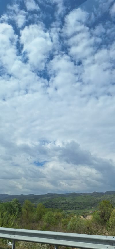 The image shows a view from a vehicle in Strumyani, Bulgaria. The sky is predominantly blue with scattered cumulus clouds. The right side of the image shows a hillside covered in green vegetation and trees, adjacent to a road with a metal guardrail.