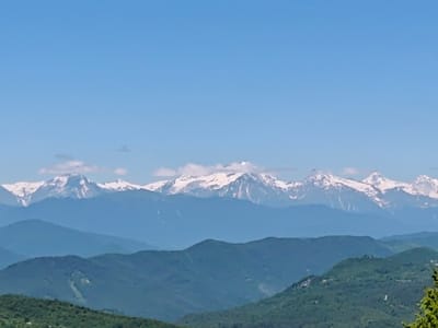 A panoramic view of layered mountain ranges under a clear blue sky. The farthest range features majestic peaks capped with snow, stark white against the sky and interspersed with fluffy white clouds. Closer mountains are progressively darker shades of blue and green, indicating increasing distance and atmospheric haze. The closest mountains in the foreground are densely covered in lush green vegetation, suggesting a temperate climate. There are no people, objects, or text visible in the image. The scene depicts a serene, natural landscape, likely captured during the daytime under fair weather conditions, given the bright sunlight illuminating the scene and the clear sky. The setting appears to be a vast, mountainous region, possibly from a high vantage point, offering a sweeping vista of the natural landscape. The overall impression is one of natural beauty and tranquility.