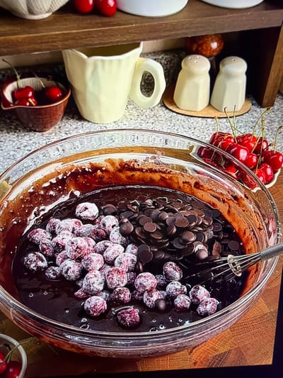 An overhead, close-up shot captures a vibrant arrangement of baking ingredients in a kitchen setting. The central focus is a large, clear glass mixing bowl containing a dark chocolate batter. Floating within the batter are numerous dark chocolate chips and a generous pile of fresh red cherries, which appear to be coated in a light dusting of white powder, likely flour or sugar, prepared for baking. A silver metal whisk rests partially submerged in the batter on the left side of the bowl.

To the left of the mixing bowl, a section of a wooden shelving unit or box is visible. It holds two off-white, faceted ceramic salt and pepper shakers, a small, reddish-brown decorative sphere, and a cream-colored ceramic pitcher with a handle. Further down, a small brown ceramic cup contains additional red cherries, and part of a white, textured object is seen in the bottom left corner.

The backdrop features a textured wooden surface, possibly a cutting board, on the right side, and a speckled grey countertop across the rest of the visible area. The scene depicts a moment of food preparation, specifically for a chocolate and cherry baked good. No individuals are present. The time of day is not discernible, and there are no specific location cues that point to Ros Na Rí, Ireland or any other geographical place. No text is visible within the image.