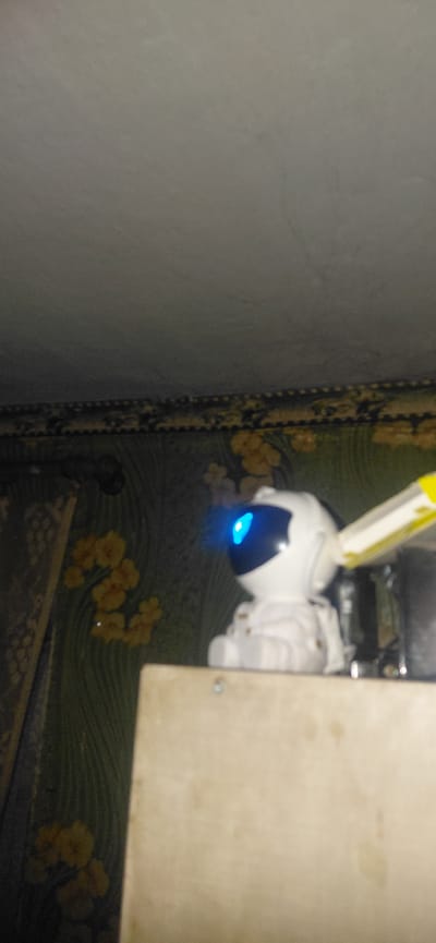 The image depicts a close-up, slightly blurry view of a white toy robot designed to resemble an astronaut, situated on a light-colored wooden surface. The robot is positioned centrally and is facing left, with a bright blue light emanating from its helmet visor. To the right of the robot, a portion of a yellow and white object, possibly another toy or a part of its accessories, is visible. The background consists of patterned wallpaper featuring green foliage and yellow flowers, suggesting an indoor domestic setting. The ceiling is visible at the top of the frame, appearing textured and uneven. The overall lighting is dim, with the blue light from the robot's visor being the most prominent light source. The location is stated to be in the city of Ketrysanivka, Ukraine.