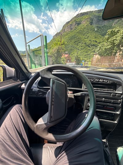 The image provides an interior view from the driver's seat of a car, looking forward through the windshield. The car's dashboard, steering wheel, and part of the driver's legs are visible. Outside the car, a paved road leads into a town with buildings, including a red-roofed structure, and a wooden fence. A steep, tree-covered mountain dominates the background, indicative of the mountainous terrain surrounding Tumanyan, Armenia. The sky is blue with scattered white clouds. The scene appears to be a sunny day.