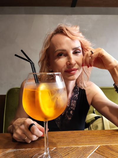 A woman with light brown hair is sitting at a wooden table with a glass of orange liquid with ice and two black straws. She is wearing a black sleeveless top with lace detailing on the neckline, a black ring with dark stones on her right index finger, and a gold ring on her left ring finger. Her right hand is resting on her cheek, and her left hand is resting on the table. She is looking at the camera and smiling. The background is blurry, but it appears to be an indoor setting with a light gray wall and a green couch. The location is Chelganovo, Russia.