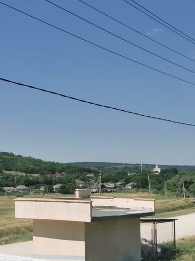 A rural landscape under a clear blue sky is depicted. In the foreground, a beige, cubic structure with a flat, tiled roof is prominently featured. A smaller, attached structure with a metal frame and wire mesh sides, possibly a shelter or enclosure, is visible to the right of the main structure. The background reveals a rolling green hillside dotted with trees and houses, suggesting a village or small town. A white building with a cross on top, likely a church, is visible in the distance on the hillside. Power lines crisscross the sky above the scene. The ground around the structures is covered in dry, yellowish grass, and a dirt road or path leads into the distance. The overall impression is of a sunny, dry day in a quiet, rural setting.