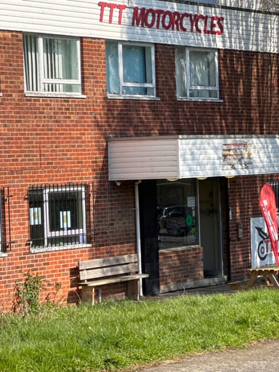 An exterior, brightly lit daytime photograph depicts a two-story commercial building located in Babergh, United Kingdom. The building features a red brick facade with white window frames and trim. The upper floor displays red lettering spelling "TTT MOTORCYCLES" on a white fascia board. Three white-framed windows are visible on the upper floor; the left window has vertical blinds, the middle has a light blue curtain or frost, and the right has a sheer curtain. All upper windows appear slightly ajar.
The ground floor features two windows secured with black metal security grates to the left of the main entrance. Posters are visible inside these windows. A weathered wooden bench with three slats is positioned on the ground immediately in front of the leftmost ground-floor window. Overgrown green grass and weeds are present near the building's base to the left of the bench.
The main entrance area is recessed and covered by a two-part awning: a light beige corrugated panel on the left and a white panel with a faded, possibly branded, graphic on the right. The entrance itself consists of a glass storefront reflecting a dark vehicle, with various stickers affixed to the glass. A dark-framed door is visible within this glass facade. A white downpipe runs vertically to the right of the entrance. To the right of the entrance, a wooden picnic table is partially visible, holding a red flag or banner with white graphics and a separate white sign. Strong shadows from unseen overhead foliage are cast across the entire building facade and the grassy foreground, indicating direct sunlight. No individuals are present in the scene. The immediate foreground is covered in unkempt green grass.