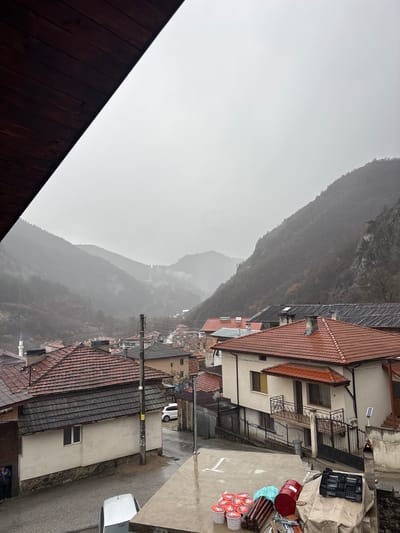 An elevated viewpoint captures a residential section of Grohotno, Bulgaria, on an overcast, possibly rainy day. The scene is dominated by a valley surrounded by steep, mist-shrouded, tree-covered mountains in the background.
In the immediate foreground, to the upper left, a dark brown wooden eaves structure is partially visible. To the lower right, a concrete ledge or balcony floor holds several items: eight red and white lidded buckets, a red metal drum, bundles of dark wooden or metal rods, a black plastic crate, and a beige tarp covering other objects.
The midground features a dense arrangement of residential buildings with various tiled roofs, predominantly red-orange and some darker grey. Most houses are light-colored (beige or white stucco). A wet asphalt street winds through the neighborhood, indicating recent precipitation. A white hatchback is partially visible on the street in the lower left, and a white SUV is parked further up the street. Multiple dark utility poles with wires run along the street.
Two individuals are visible standing near the entrance of a light-colored building with a dark-tiled roof in the lower-middle left. In the mid-right, a multi-story house with a beige facade features a prominent balcony with ornate black railings and a red-tiled awning. A small solar panel is mounted on a pole beside this house.
In the far left midground, above the rooftops, a slender white minaret with a conical roof is visible, suggesting the presence of a mosque. The overall environment conveys a mountainous village setting with a significant residential presence.