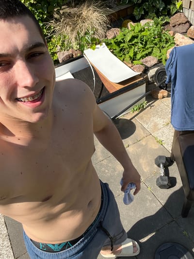 A shirtless young man wearing braces smiles at the camera. He is wearing blue pants and a white sandal. He holds a white cloth in his hand. The background includes paving stones, a weight bench, a dumbbell, weights, rocks, and foliage. The location appears to be an outdoor space, possibly a yard, but the city of Oud-Beijerland, Netherlands, cannot be confirmed.