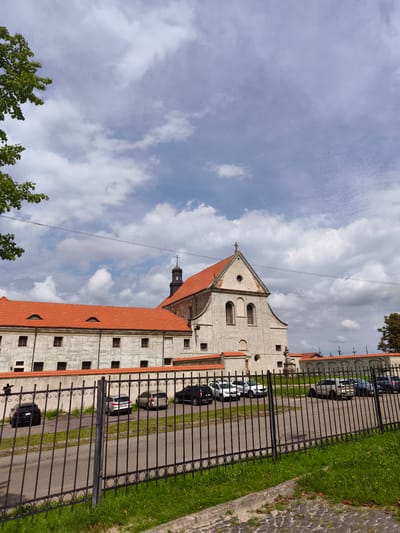 This image depicts the exterior of a historic religious building, likely a monastery or church, situated in Olesko, Ukraine. The architecture features a prominent red-tiled roof, a bell tower topped with a cross, and light-colored walls with multiple windows. A black metal fence runs along the foreground, partially obscuring a parking lot where several cars are parked. Lush green grass and a paved area with cobblestones are also visible around the building and fence. The sky above is partly cloudy with a mix of white and grey clouds, suggesting moderate weather conditions.