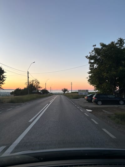 The image shows a road in the outskirts of Pașcani, Romania, at dusk. The sky is a gradient of pale blue at the top, transitioning to orange and pink hues near the horizon, indicating either sunrise or sunset. A paved road with double white lines runs through the center of the frame, extending into the distance. On the left side of the road, there are grassy fields, trees, and utility poles with overhead wires. In the distance, a town or city can be seen, partially obscured by a misty layer close to the horizon. On the right side of the road, there is a large tree, a signpost with some text, and two parked cars. The foreground shows the dashboard or hood of a vehicle, suggesting the image was taken from inside a car. The overall atmosphere is serene and tranquil.