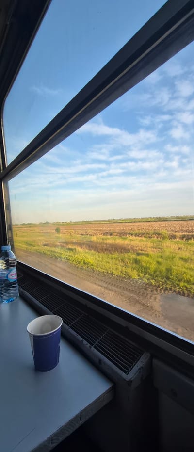 The image presents a view from inside a moving vehicle, likely a train or bus, looking outwards through a large, possibly panoramic, window or skylight. The scene is captured with a tilted perspective, causing the horizon line of the landscape to appear almost vertical.

Outside, a wide expanse of rural or agricultural land unfolds under a bright blue sky adorned with scattered, wispy white clouds. The landscape consists of various fields, some vibrantly green with growing crops, and others appearing recently tilled or fallow, displaying earthy brown tones. These fields suggest a setting in an agricultural region, aligning with the provided location context of Manolsko Konare, Bulgaria.

Inside the vehicle, on a surface adjacent to the window in the lower right, a clear plastic bottle of water is visible. The label on the bottle prominently features the brand name "БАЛДЕРАН" (Balderan) and the text "НАТУРАЛНА МИНЕРАЛНА ВОДА" (Natural Mineral Water). Next to the bottle is a dark blue disposable paper cup with a white interior and rim. A metal grate, resembling an air vent, is also part of the vehicle's interior structure, along with dark paneling and a lighter grey surface.

The bright natural light indicates it is daytime with clear, fair weather conditions. The implied movement of the vehicle suggests a journey, offering a tranquil observation of the passing countryside. No people are visible in the frame.