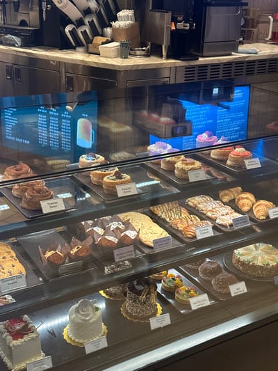 The image depicts the interior of a bakery or cafe in Paradise, United States, showcasing a tiered glass display case filled with various baked goods and desserts. No individuals are visible within the scene.
The upper portion of the image features a white marble-patterned countertop equipped with commercial coffee machines, stacks of disposable white coffee cups and lids, a box of white straws, a pile of white paper napkins, and a dark green ceramic mug. Below this counter, stainless steel cabinetry is present.
The prominent glass display case contains three shelves of items.
On the top shelf, various individual pastries are arranged on dark rectangular trays, including "Tiramisu Donut," "Pistachio Donuts," a "Latte Pie Donut," and a "Strawberry Shortcake Donut."
The middle shelf holds larger baked goods, such as a large "Chocolate Chip Cookie," "Peanut Butter Cookie (Gluten Free)," "Blueberry Muffin (gluten free)" in paper liners, "Chocolate Croissant," "Fruit Danish/Cheese Figuri," and plain croissants. Each item is individually presented with identifying labels.
The bottom shelf displays small cakes and tarts, including a square "Strawberry Shortcake," a round "Celebration Vanilla" cake adorned with a white rose, a "Celebration Chocolate" cake, "Green Tea Custard Fruit Tart" pastries, and several "Mini Bundt Cake" items. These items also have price or description labels.
Behind the display case, two vertical digital screens with a blue hue are visible, displaying text that appears to be menu listings or advertisements, alongside internal reflections. The overall scene indicates a clean, organized, and well-stocked food service establishment.