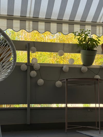 This media file captures an outdoor scene, likely on a sunny day. The primary focus is a decorative string of white paper lanterns, illuminated and hanging against a backdrop of lush green foliage. To the left, a striped awning in shades of grey and white provides shade, with a scalloped edge adding a decorative touch. A potted plant with vibrant green leaves is positioned on a shelf, adding to the natural element of the scene.
The composition suggests a relaxed, outdoor setting, possibly a patio or garden area. The bright sunlight filtering through the leaves indicates daytime. The overall mood is serene and pleasant, enhanced by the gentle glow of the lanterns. The image also features parts of outdoor furniture, including the top of a white wicker chair in the foreground, suggesting a place for leisure and relaxation. The scene is framed by vertical structural elements, possibly part of a porch or pergola, and a glimpse of a rug with a blue patterned border is visible on the right. No text is visible in the image.
