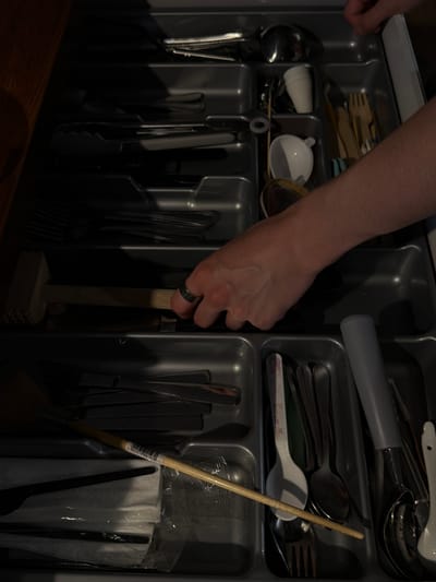 The image shows an open kitchen drawer filled with various utensils. A person's hand, wearing a silver ring, is visible reaching into the drawer. The hand is grasping the wooden handle of what appears to be a meat mallet, which is positioned vertically within the drawer. The drawer organizer is made of grey plastic and is divided into compartments.

In the upper compartments, several spoons and forks are neatly arranged. To the left, a ladle and other larger utensils are stored. In the middle section, there are white and blue items, possibly lids or small containers, alongside more cutlery. Further to the right, a long wooden stick, possibly a stirrer or a tool for baking, with a barcode sticker attached, is leaning against other items.

The scene is indoors, likely a kitchen, given the presence of kitchenware. The lighting suggests it might be daytime or artificial light is being used, casting shadows within the drawer. The overall impression is one of organized storage of kitchen tools. There is no visible text other than the barcode sticker on the wooden stick. The location context of Słupsk, Poland, does not provide specific visual cues within the image itself.