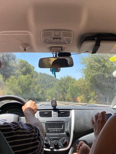 The image depicts the interior of a vehicle, viewed from the front passenger seat, traveling on a paved road in Mas'ade, Israel. A male driver, wearing a striped shirt, has both hands on the steering wheel, indicating active operation of the vehicle. To the right of the driver, bare feet are visible, presumably belonging to the front passenger, resting on or near the dashboard area. A dark-colored phone mount is affixed to the center dashboard. The car's dashboard features a radio unit and climate controls. The rearview mirror reflects a partial view of an individual's head and shoulders, likely the passenger, and a hanging blue beaded decoration. The windshield appears to have accumulated dirt and smudges. Outside the vehicle, a clear blue sky is visible above a landscape dominated by dense green foliage and trees, indicative of hills or mountainous terrain. A winding road extends into the distance, with at least two other vehicles observable ahead. A light-colored sun visor, with an attached dark strap and pouch, is partially lowered on the passenger side. A yellow sticker with Hebrew text is present on the passenger-side windshield, near the sun visor.