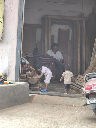 The image shows an outdoor scene in Malkapur, India, likely in front of a shop. In the foreground, on the left, is a concrete structure with some marigolds and green leaves placed on it, suggesting a small shrine. Adjacent to this is a pile of cut wooden logs with orange electrical wires draped over them. A young child, wearing a light pink t-shirt and shorts, is crouched down, appearing to interact with the logs and holding a red object, possibly a marker or crayon. To the right, a younger child, wearing a beige t-shirt and patterned shorts, stands with their back to the camera. In the background, inside a dimly lit doorway, a man is seated in a chair, looking to the right. The doorway is filled with stacked wooden frames and planks, indicating a carpentry or woodworking establishment. A ceiling fan is visible in the upper background. On the far right of the image, the rear of a scooter with a license plate reading "MH28B S7773" is partially visible. The ground is a mixture of dirt and some paving.