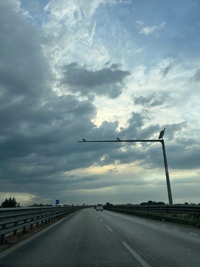 The image shows a view from a vehicle driving on a highway in Barletta, Italy, under a cloudy sky. The road is paved and has white lane markings. Guardrails line the left side of the road, and a pole with a solar panel and what appears to be a camera or sensor is visible on the right side. Several other vehicles, including cars and vans, are on the highway ahead. The surrounding landscape is mostly flat with some vegetation and utility poles in the distance. The sky is dramatic with dark storm clouds mixed with lighter patches of sky where sunlight is breaking through.