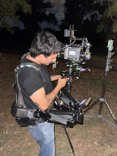 A cameraman, wearing a camera harness, operates a professional camera mounted on a portable rig. The man wears a black shirt, blue jeans, and eyeglasses. The background is a dimly lit outdoor area, possibly a wooded area, with trees visible in the distance. Additional film equipment, including stands, are present. The camera rig itself is complex, with various accessories and monitors attached. The scene suggests a film or video production taking place at night.