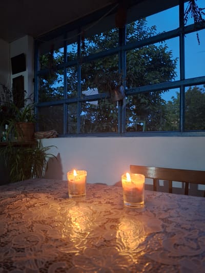 This is a medium shot, horizontal photograph taken at dusk or dawn in Nabran, Azerbaijan. The image captures an intimate and serene scene set on a table covered with a white lace tablecloth. Two lit candles in clear glass holders are placed on the tablecloth, casting a warm, flickering glow and creating dancing shadows on the intricate lace pattern. The light from the candles illuminates the immediate foreground.
Beyond the table, a large window or glass roof structure is visible, divided by dark blue framing. Through the glass, the twilight sky is a deep, clear blue, suggesting either early morning or late evening. Silhouetted against the sky are the dark shapes of trees and foliage, their leaves creating a textured pattern. The overall mood is tranquil and peaceful, evoking a sense of calm and quiet reflection. The setting appears to be indoors or a covered outdoor space, looking out onto a natural landscape during the transition from day to night or vice versa. There is no visible text in the image.