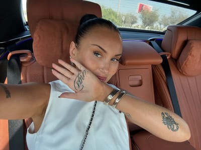 A female individual is depicted taking a selfie from the rear passenger seat of a luxury vehicle, located in Al Bahyah, United Arab Emirates. She has dark hair pulled back in a bun, blue eyes, and is wearing a white sleeveless top. Her right hand is raised, palm facing the camera, partially obscuring her lower face. A "virgo" tattoo is visible on the back of her right hand, and a scorpion tattoo is present on her left forearm. Two ribbed metal bangles adorn her left wrist. The vehicle's interior features brown leather upholstery on the seats, headrests, and a central console. Blue ambient lighting is visible on a door panel. Through the rear window, a sunny outdoor environment is visible, showing sparse green trees, a road, and a distant billboard depicting a female figure on a red background.