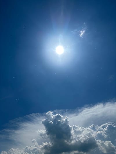 This media file captures a bright, sunny day with dramatic cloud formations in the sky. The sun is a prominent, luminous orb in the upper left quadrant, casting a bright glare and visible rays. Scattered across the deep blue sky are numerous small, white specks that could be dust, pollen, or tiny insects. To the right, a large, dynamic cloud bank stretches vertically. The clouds are a mix of bright white where the sun hits them directly and darker, shadowed grays in their denser sections. The contrast between the clear blue sky and the textured, voluminous clouds suggests a partly cloudy day with potential for changing weather.

The image is oriented vertically, making the cloud formation appear as a towering wall. The scene is devoid of any people or man-made objects, focusing solely on the natural elements of the sky. The bright sunlight and the presence of sunlit clouds indicate it is likely daytime, possibly late morning or early afternoon. The overall mood is one of natural grandeur and the power of atmospheric phenomena. No text is visible in the image. The location context provided is Pravda, Russia, which, combined with the visual cues, paints a picture of an outdoor scene in that region on a bright, potentially warm day.