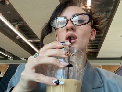 The image captures a close-up, upward-angled view of an individual, likely female, consuming a beverage from a glass through a straw in an indoor setting within Garston, United Kingdom. The subject's face is partially visible, dominated by oval, tortoiseshell-patterned eyeglasses reflecting interior lights and a distant outdoor scene. A small hoop nose ring is present on the left nostril, and multiple small piercings adorn the visible right earlobe and helix. Dark hair is pulled back, with some strands appearing disheveled. The subject's lips are parted around a black straw inserted into a clear glass. The left hand, positioned centrally, holds the glass. This hand features a silver-toned ring with a discernible emblem on the index finger. The fingernails are long, artificial, and display varied designs including black French tips, black polka dots on a nude base, and black-and-white abstract patterns. The glass contains a light brown, opaque liquid. The individual is wearing a grey blazer or jacket, with a slender chain necklace visible. The background reveals an indoor environment with an exposed ceiling featuring prominent linear LED light fixtures and visible structural or ductwork elements, suggesting a commercial or institutional space.