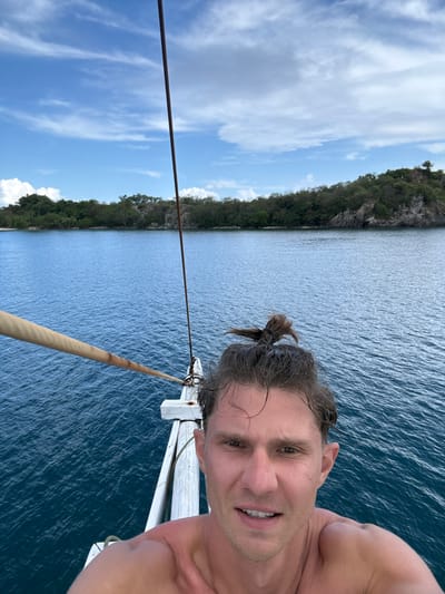 The image shows a selfie of a fair-skinned man with wet hair pulled back into a small ponytail. He is smiling slightly at the camera. The background features a body of water, possibly a bay or lagoon, with land on the opposite side covered in dense green vegetation. The sky shows patchy clouds. The man appears to be on a boat, as indicated by the white railings and ropes in the foreground. The location is likely in the vicinity of Pasir Putih, Indonesia given the water and land formations.
