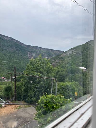 A view from a window shows a mountainous landscape covered in lush green trees under a cloudy sky. A cluster of buildings, including a red-roofed house, is visible nestled at the base of the mountains. Utility poles and wires crisscross the foreground. A small, light-colored house with a white roof is perched on the hillside, further up the mountain. The scene appears to be in or around Tumanyan, Armenia, given the characteristic mountainous terrain and architecture. The overall impression is one of a rural, somewhat rustic environment.