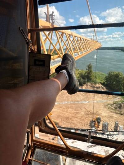 This first-person perspective photo shows a person's leg and foot resting on the controls of a crane cabin. The leg is extended outwards, with a dark sneaker and white socks visible. The background through the window reveals a construction site with a large yellow crane, a river, trees, and a blue sky with white clouds. Construction equipment, including a yellow excavator, can be seen on the dusty ground. The scene suggests a working environment, likely during the daytime given the bright sunlight and shadows. A chart with text in Polish and English, likely a load capacity chart for the crane, is mounted on the control panel. The text includes information like "Tragfähigkeit," "Capacity," "Charge," "Ausladung," "Radius," and "Portée," along with numerical values for radius in meters and capacity in kilograms. The context suggests this is a photo taken by a crane operator in Mikołajki, Poland.