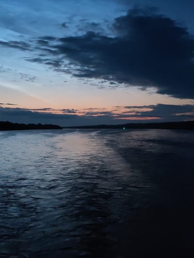 The image shows a wide body of water, likely a river or lake, with a dark, tree-lined shore in the distance. The sky above is a deep twilight blue with dramatic, dark clouds overhead, transitioning to lighter hues of pink and orange near the horizon. The water's surface is rippled and reflects the dim light of the sky, with a faint trail of disturbed water extending from the foreground into the distance. A small, bright green light is visible on the water near the distant shore. The scene appears to be a natural landscape at dusk or dawn. There are no people or man-made structures other than the distant green light. The location is identified as Ankimarovo, Russia.