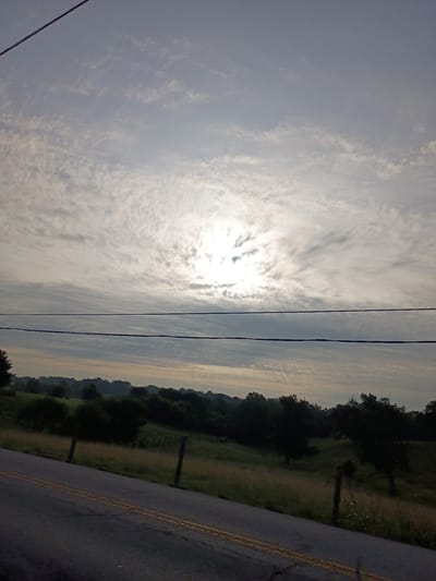The image depicts a rural landscape scene with a road running along the right side, separating it from a field. The sky is filled with scattered clouds, and the sun is partially visible, suggesting it is either sunrise or sunset. The time of day appears to be early morning or late evening, given the lighting conditions. The weather seems to be partly cloudy. The scene is set in a natural environment with trees and greenery visible in the distance. There are also power lines crossing the sky, indicating the presence of infrastructure. The overall mood is serene and tranquil. The image is oriented vertically, with the sky occupying the left half and the landscape and road on the right. There are no people or animals visible in the image, nor any discernable text.