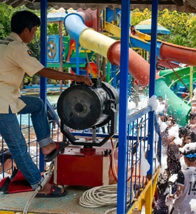 A man stands on a platform, operating a large foam machine at the Hairos Water Park in Durin Simbelang, Indonesia. He wears a short-sleeved beige shirt, blue jeans, and sandals, and is positioned next to the machine, which is on top of a red box. The machine sprays foam onto a crowd of people gathered below. Water slides in bright colors are visible in the background. A coiled white hose rests on the platform near the machine.