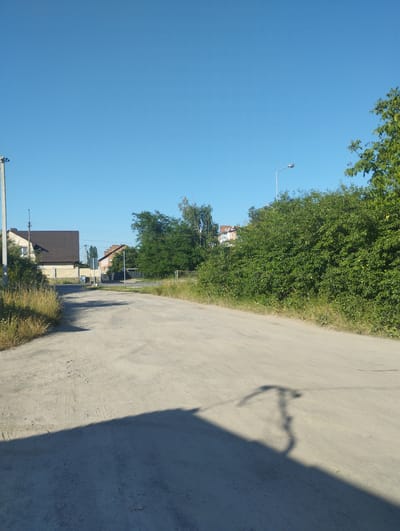 The image depicts a rural street scene in Hnivan, Ukraine. A dirt road curves through the frame, with dry grass and vegetation lining its edges. On the left, a house with a brown roof and a beige brick facade is visible. Further down the road, another building with a brick and white facade is partially obscured by trees. A tall street lamp stands on the right side of the road, with a shadow cast on the dirt path. Lush green trees and bushes dominate the right side of the image, creating a dense natural barrier. The sky is clear and blue, indicating fair weather. The sun is casting long shadows, suggesting it is either early morning or late afternoon. There is no discernible text in the image. The overall impression is a quiet, serene, and somewhat undeveloped rural setting.