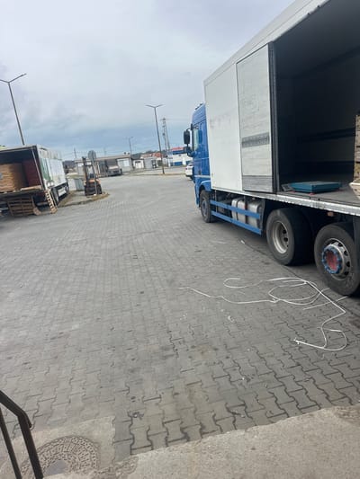The image depicts an outdoor scene in what appears to be a loading area or parking lot with gray brick pavement. A semi-trailer truck, blue and white, is parked on the left side of the frame with its cargo door open. It contains wooden pallets and material that may be packed potatoes, onions or other product. A forklift, situated behind the trailer, is visible in the distance. Several other trailers, container units and transport trucks are parked further down the open lot. There are lampposts and other structures in the background. White, possibly discarded plastic strapping lies on the pavement. The sky is overcast. The location is presumed to be in the city of Plodovitovo, Bulgaria.