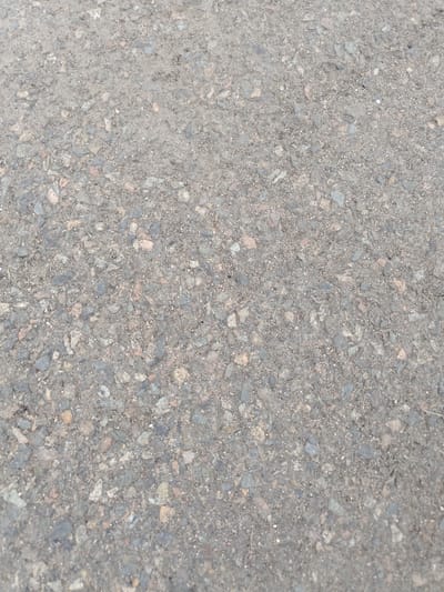 The image shows a close-up, overhead view of asphalt pavement. The surface is composed of small grey, black, and reddish-brown aggregate mixed with a dark binder. Some larger, irregularly shaped stones are visible within the asphalt. There are no people, structures, or distinct objects present in the frame. The lighting suggests an outdoor setting, likely during daylight hours. The image's context places it in the city of Stepnoe, Russia, though no specific landmarks or environmental features are identifiable within the image itself to confirm this location.