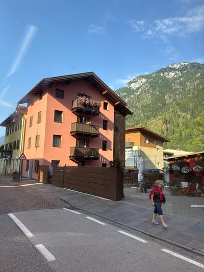 The image shows a street scene in Predazzo, Italy. A woman with gray hair wearing a red scarf over a black shirt and blue capri pants walks on a sidewalk. She is carrying a black bag. The street is paved with asphalt and has white lane markings. A cobblestone sidewalk runs parallel to the street, and beyond that, a paved walkway with a wooden fence. Buildings line the street, including a salmon-colored building with balconies adorned with flowers and a yellow building with shutters. In the background, a steep, forested mountain rises under a clear blue sky with some wispy clouds. A traffic sign depicting children crossing is visible near the yellow building. The overall scene suggests a quiet day in a town situated in a mountainous region.