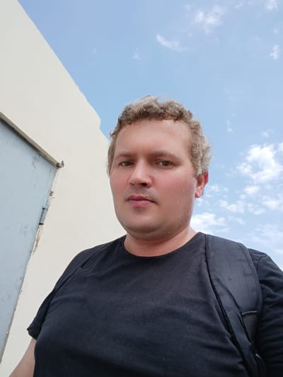 A man with curly blond hair, wearing a black t-shirt and a black backpack, stands against a background of a light-colored building and a blue sky with clouds. The man is looking directly at the camera, and his face appears slightly sweaty. The building on the left has a gray metal door. The image is a selfie taken outdoors. The location is Buzovna, Azerbaijan.