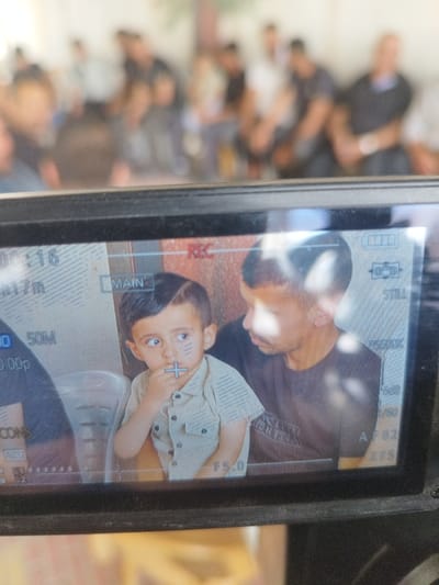 This image depicts the screen of a black digital video camera, showcasing a scene within the city of Idhna, Palestinian Territory. The primary subject displayed on the screen is a young boy with dark hair, wearing a light-colored, striped, short-sleeved button-up shirt. He has black markings on his face: two horizontal lines on each cheek and a plus sign on his chin. His right hand is positioned near his mouth, and he is looking intently to his left. Behind and to the right of the boy is an adult male with short dark hair and a dark short-sleeved shirt, whose gaze is directed towards the boy. A reddish-brown wall is visible in the background behind the two individuals, and a portion of a light-colored chair is present under the boy. The camera screen displays various digital overlays, including "REC" in red, "MAIN", "00:16" (timecode), "50M", "1080p", "F5.0", "AF82", and a battery indicator. The foreground consists of the camera's black casing, while the background beyond the camera is heavily blurred, revealing multiple indistinct human figures gathered in what appears to be an indoor or sheltered environment.