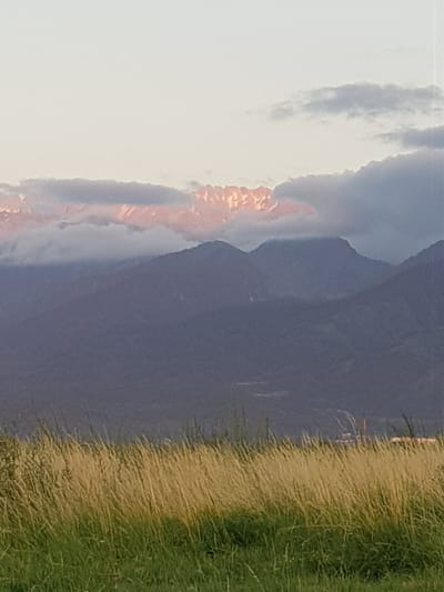 In a wide shot of a landscape, the foreground features a field of tall, dry grass with some green blades at the bottom. The midground shows dark, hazy mountain ranges layered behind one another. The background is dominated by a pale sky with scattered, wispy clouds. Towards the top of the mountains, a band of pinkish-orange light illuminates snow-capped peaks, partially obscured by clouds. In the far distance, on the right side of the frame, a glimpse of industrial structures, possibly a factory with a chimney, can be seen through the hazy mountains. The scene appears to be taken during either sunrise or sunset, given the warm light on the mountain peaks.