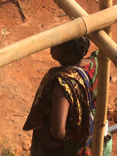 In Agukwu Nri, Nigeria, a partial view of an individual is observed from behind in an outdoor setting. The person has dark, braided hair pulled back and is wearing a colorful, patterned fabric wrap in shades of red, green, blue, yellow, and black. A bare, dark-skinned arm is visible. The individual is positioned over uneven, reddish-brown earth, characteristic of laterite soil. Partially obscuring the scene are two light brown bamboo poles, one horizontal and one vertical, forming a cross. These poles exhibit visible nodes and small dark blemishes. A small portion of a green object or container and a dark pipe are discernable in the lower right, behind the vertical bamboo pole. Bright daylight illuminates the scene.