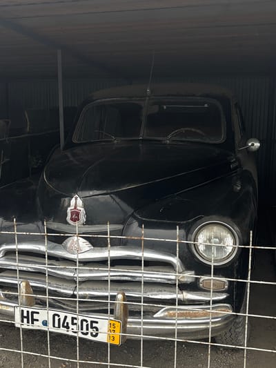 The image shows the front of a vintage black car, possibly a GAZ-21 Volga, parked inside a garage or shed. The car is dark and dusty, with chrome accents on the grille, headlights, and bumper. A license plate with the characters "HF 04505" is visible on the front bumper, along with smaller yellow plates. The car is partially obscured by a wire fence. The surroundings are dark and appear to be industrial or utilitarian. The location is stated to be Bol'shoi Mashlyak, Russia.