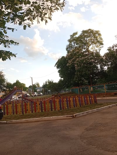 The image depicts a playground in Rezina, Moldova, likely on a warm day given the presence of leafy trees and a bright sky. In the foreground, a colorful wooden fence with alternating purple, yellow, and red posts separates the playground from a paved area. Behind the fence, a children's playground structure with a slide and swings is visible, along with grass and some dirt. Several cars are parked in the background, suggesting the playground might be near a residential area or a public space. The sky is partly cloudy with bright sunlight, casting shadows and illuminating the scene. A large tree with white blossoms stands prominently in the background, adding to the natural beauty of the location. The overall impression is a calm, everyday scene in a small town setting.