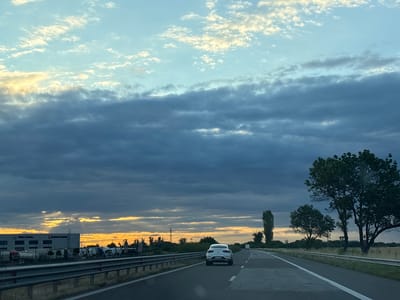 The image depicts a view from a vehicle traveling on a highway, presumably near Benkovski, Bulgaria. The sky is a prominent feature, with layers of clouds, some dark and heavy, others lighter and tinged with yellow from the setting or rising sun. Shafts of light break through the darker clouds on the left side of the frame, illuminating the horizon.
On the left side of the highway, a large industrial building is visible, with the text "Volvo Trucks" clearly displayed on its facade. Several trucks and trailers are parked in front of the building. The highway itself is paved with asphalt, marked with white lines indicating lanes. A metal guardrail runs along the side of the road.
In the foreground, a white car is driving ahead on the highway. Further down the road, more vehicles and trees are visible. The landscape on the right side of the highway consists of fields and trees, including several tall, slender trees and a larger, fuller-crowned tree. The overall impression is a highway scene at dusk or dawn, with dramatic cloud formations and industrial elements on one side, and natural landscape on the other.
