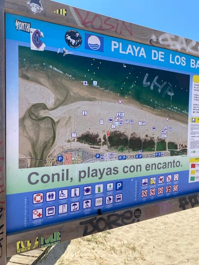 The image depicts a large informational sign located on a beach in Conil de la Frontera, Spain. The sign is mounted on a wooden frame and is primarily blue and green. At the top, the sign displays "PLAYA DE LOS BA" in white lettering, suggesting it is part of a title related to a beach. Below this, a map of the beach area is visible, showing the coastline, sandy areas, and some vegetation. Several small icons are scattered across the map, indicating various amenities or zones.
The lower portion of the sign features text and icons. A large green band across the bottom reads "Conil, playas con encanto." Below this, there are two columns of icons with corresponding text in Spanish. The left column, labeled "INSTALACIONES FACILIDADES," includes icons for a lifeguard, restrooms, accessible restrooms, controlled beach access, food and drink service, and sports zones. The right column, labeled "CONDUCTA," displays icons related to beach regulations, such as no campfires, no horses, no dogs, no tents, and no vehicles.
The sign itself shows signs of wear and tear, with stickers and graffiti applied to it. Notably, there are several stickers with graphics and text, including one with a monkey-like figure and the word "MONTHI." Graffiti in red and white spray paint is visible on the wooden frame and the blue section of the sign. The background of the image shows a sandy beach with the ocean in the distance, indicating it is an outdoor, sunny environment. The overall scene is an informative public display on a beach, subject to the elements and public interaction.