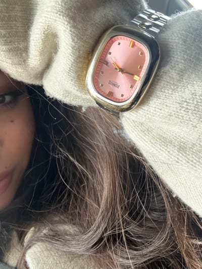 A close-up image, set in Luc-sur-Orbieu, France, features the partial left profile of an individual. Visible elements include their left eye, a section of their mouth, and abundant dark brown, wavy hair which occupies the lower half of the frame. The individual is partially obscured by a cream-colored, textured garment, possibly knitted, which wraps around their head and neck area. Prominently displayed in the upper right is a gold-toned, rectangular Ronius Precision wristwatch with rounded corners. The watch face features a salmon-pink dial with gold hour markers, gold hour, minute, and second hands. A date window at the 3 o'clock position shows the numeral '8'. The watch is affixed to a silver-toned metal link bracelet. The immediate background is out of focus and indistinct, providing no specific environmental details beyond general ambient lighting.