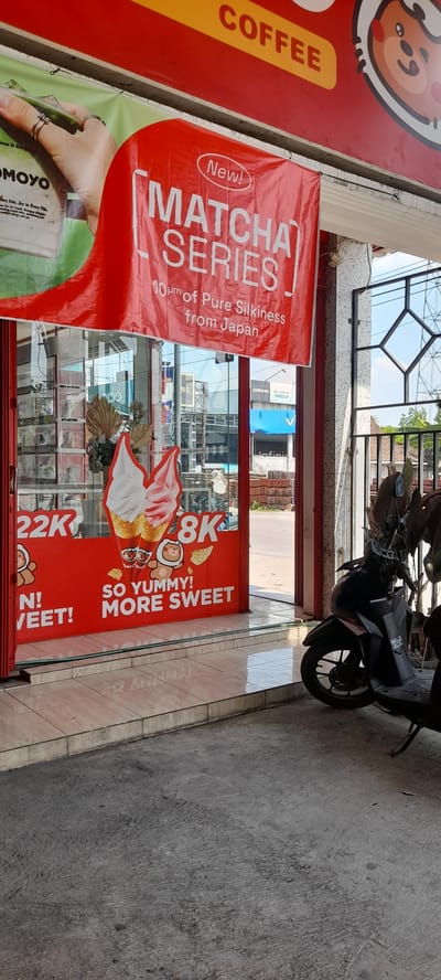 In Crewek, Indonesia, the image captures the exterior of a storefront featuring a prominent red banner advertising a "Matcha Series" and a "Coffee" section at the top. The storefront has large glass doors revealing an interior with displays. On the glass doors, there are large graphics of ice cream cones and text indicating prices, such as "22K" and "8K", along with the slogan "SO YUMMY! MORE SWEET!". To the right of the entrance, a black motorcycle is parked. The surrounding environment shows a street with buildings and vegetation. The lighting suggests it is daytime.
