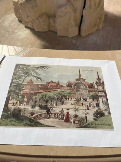 The image is a vintage illustration depicting a bustling scene in Évian-les-Bains, France. In the background, an ornate building with multiple domes, spires, and a prominent clock tower is visible. The building appears to be a grand establishment, possibly a casino or thermal bath house, with large arched windows and intricate architectural details. The foreground shows a manicured garden with trees, pathways, and people engaged in various activities. Several individuals are dressed in period clothing, with men in suits and hats, and women in long dresses. Some are walking, others are conversing, and a horse-drawn carriage is present, suggesting a leisurely atmosphere. A sign in the upper right corner reads "EVIAN-LES-BAINS," and a smaller inscription at the bottom right states "Buvette de la Source Cachat." The overall impression is one of a lively, upscale resort town during a bygone era.